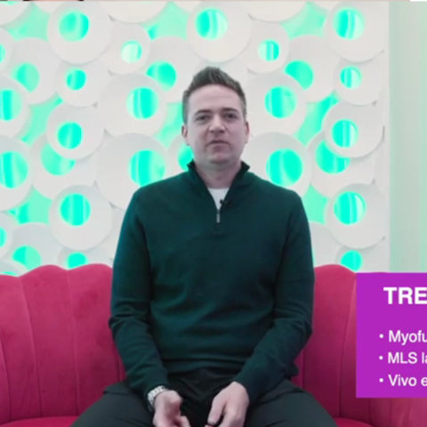 A man in a dark sweater sits on a red couch in front of a white patterned wall with teal lighting. A purple box with text appears in the bottom right corner.
