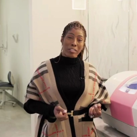 A woman stands indoors next to a pink and white medical or cosmetic device, wearing a black top and a patterned shawl.