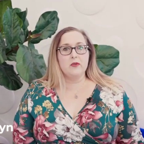A woman with long light brown hair and glasses, wearing a floral dress, sits in front of a large leafy plant and a white wall.