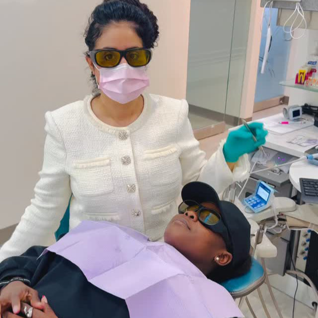 A dentist wearing protective glasses and a mask stands beside a patient seated in a dental chair, both wearing protective eyewear in a dental clinic.