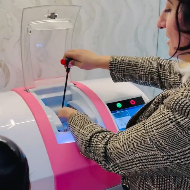 A person in a plaid jacket operates a pink and white laboratory machine, using a long tool to adjust or insert something inside the open compartment.