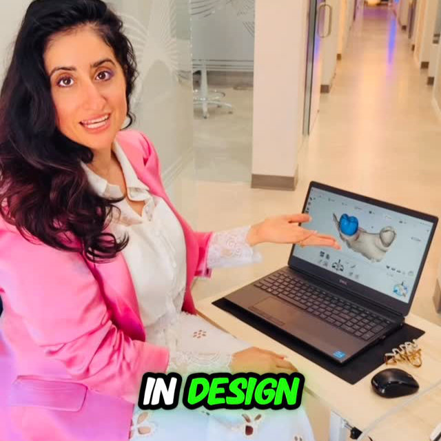 A woman in a pink blazer sits at a desk in an office hallway, gesturing to a laptop displaying a 3D hand design on the screen. Text overlay reads "IN DESIGN.