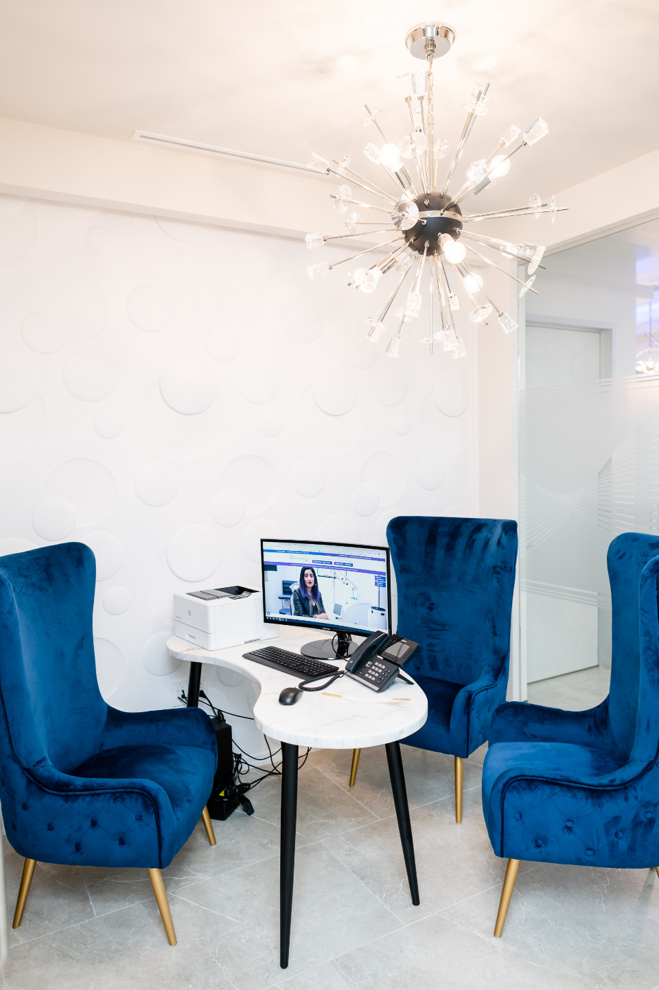 A small office area with a white desk, three blue velvet chairs, a computer monitor, phones, and a modern chandelier overhead.