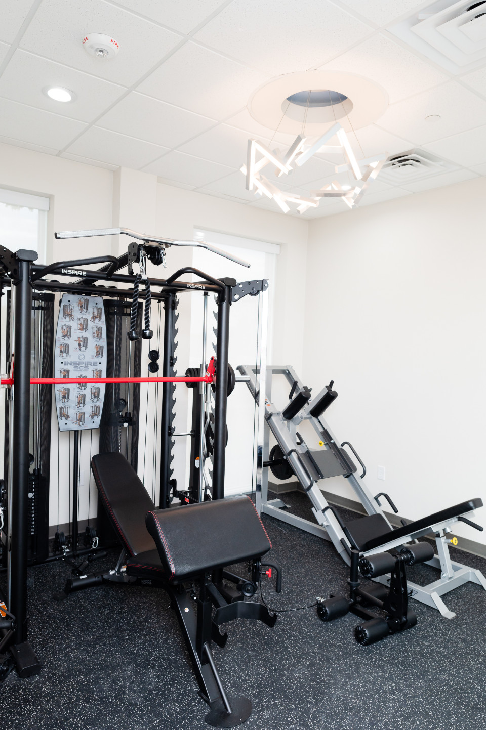 A small gym room with a weight machine, a bench press, a leg press machine, and black rubber flooring under bright ceiling lights.