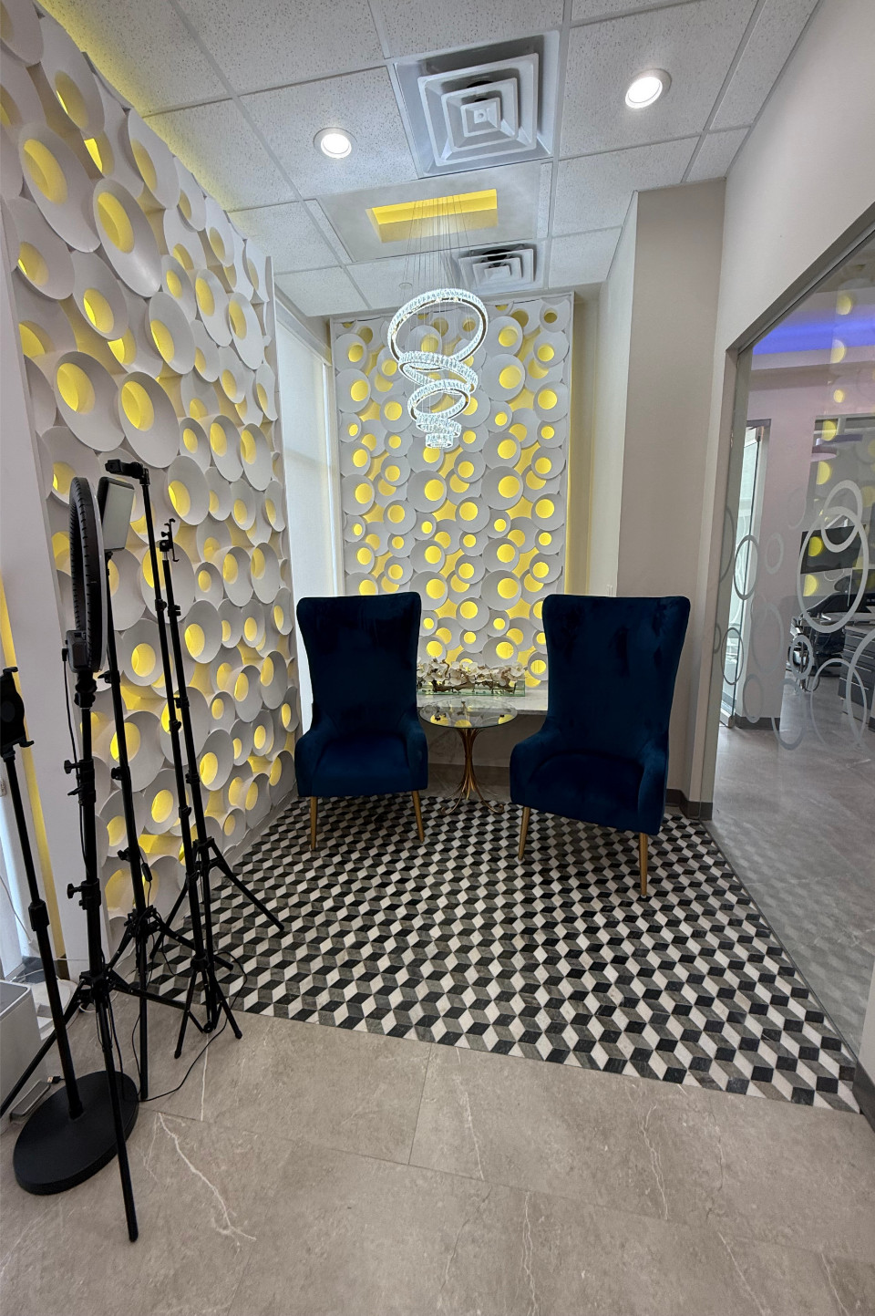 Two blue velvet chairs and a small table sit in front of a decorative wall with circular cutouts and yellow lighting; photography equipment is on the left.
