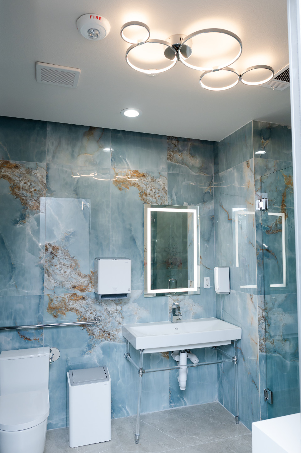 Modern bathroom with blue marble-patterned walls, white fixtures including sink and toilet, a mirror with built-in lighting, ceiling light, and a small trash bin.
