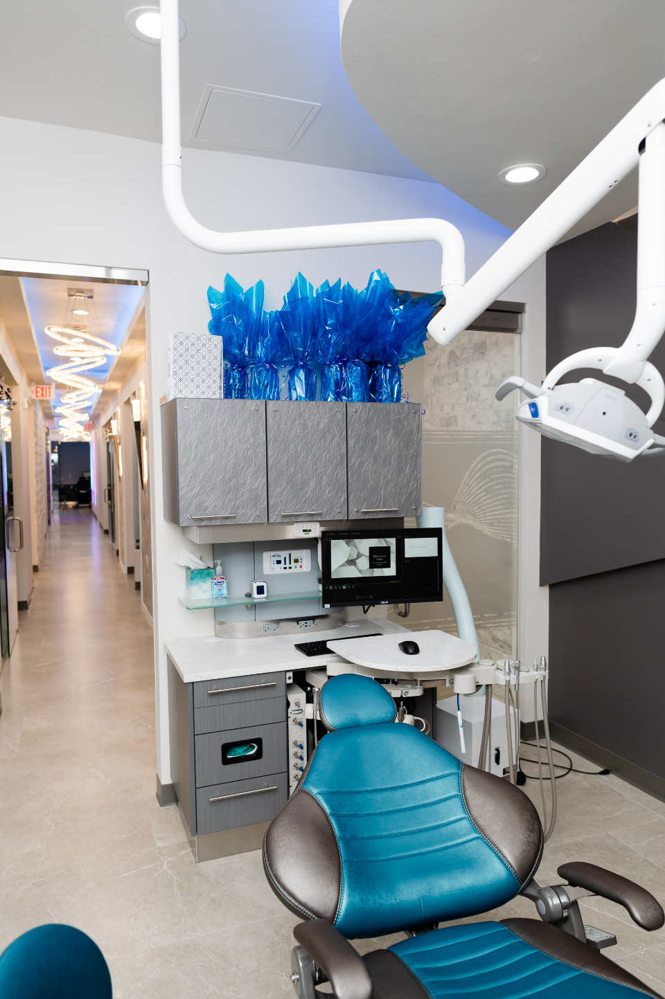 A modern dental exam room with a blue dental chair, equipment, cabinets with blue-wrapped items on top, and a hallway visible in the background.