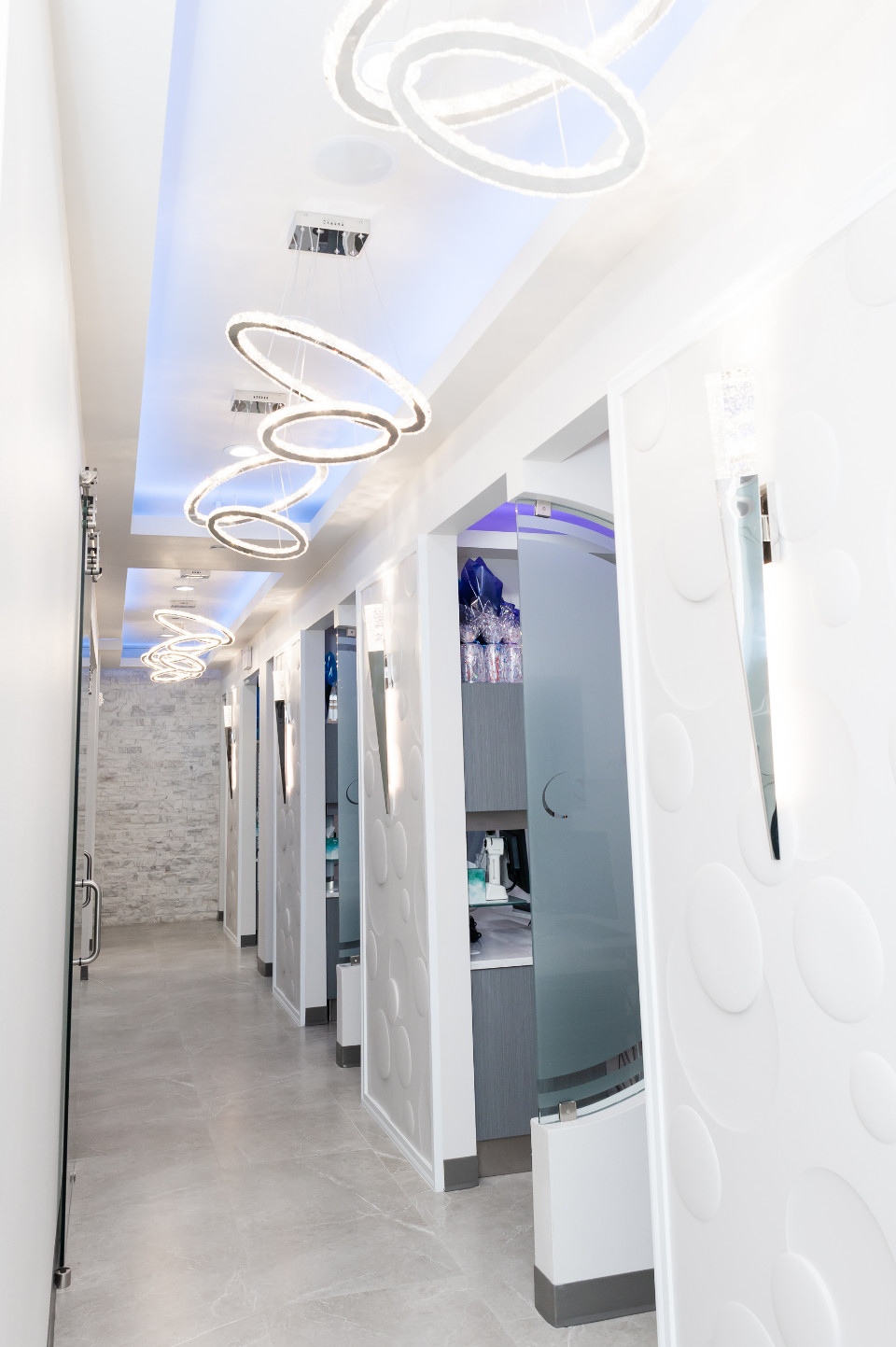 A modern clinic hallway with circular light fixtures, white walls, and glass-partitioned rooms, leading to a stone-textured wall at the end.
