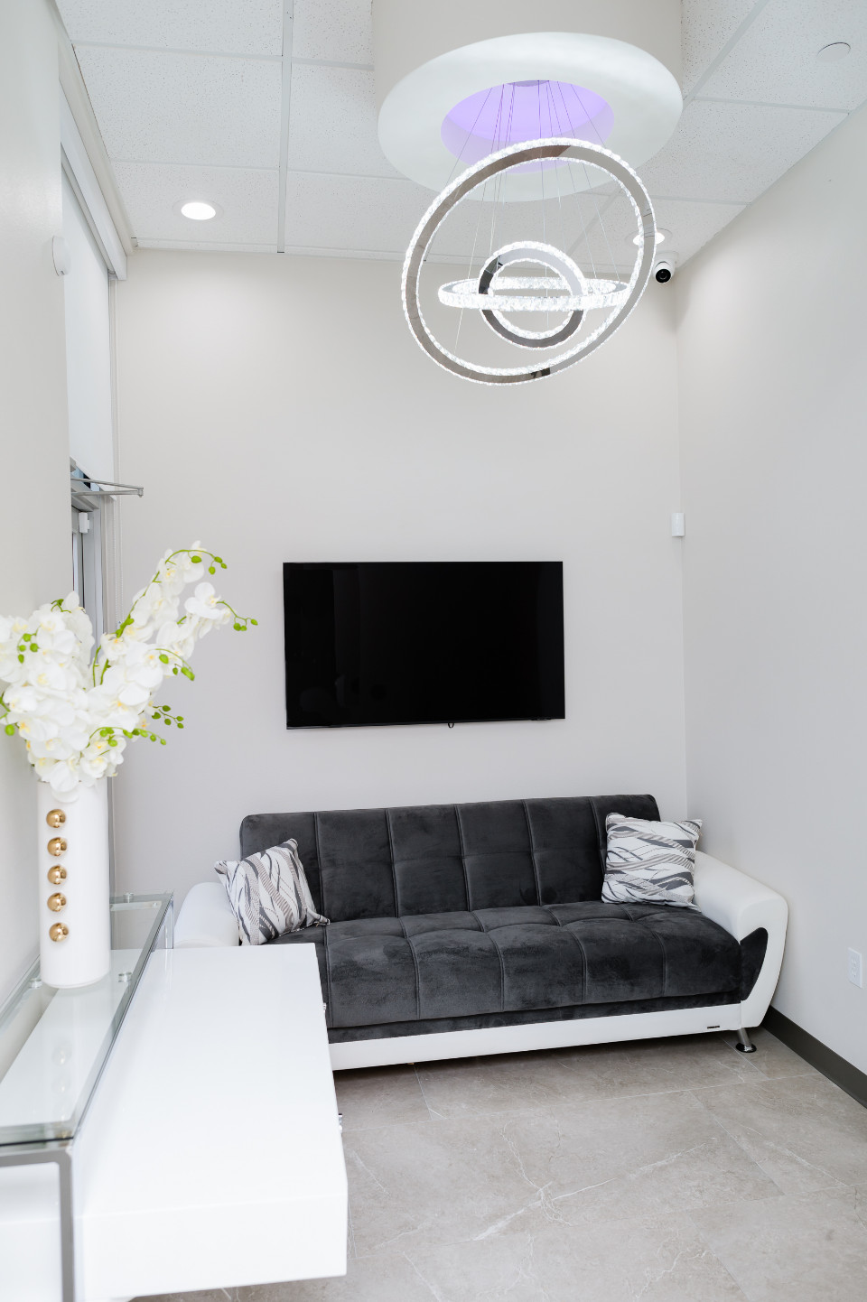 A modern waiting room with a gray and white sofa, a wall-mounted TV, a glass table with white flowers, and a circular ceiling light fixture.