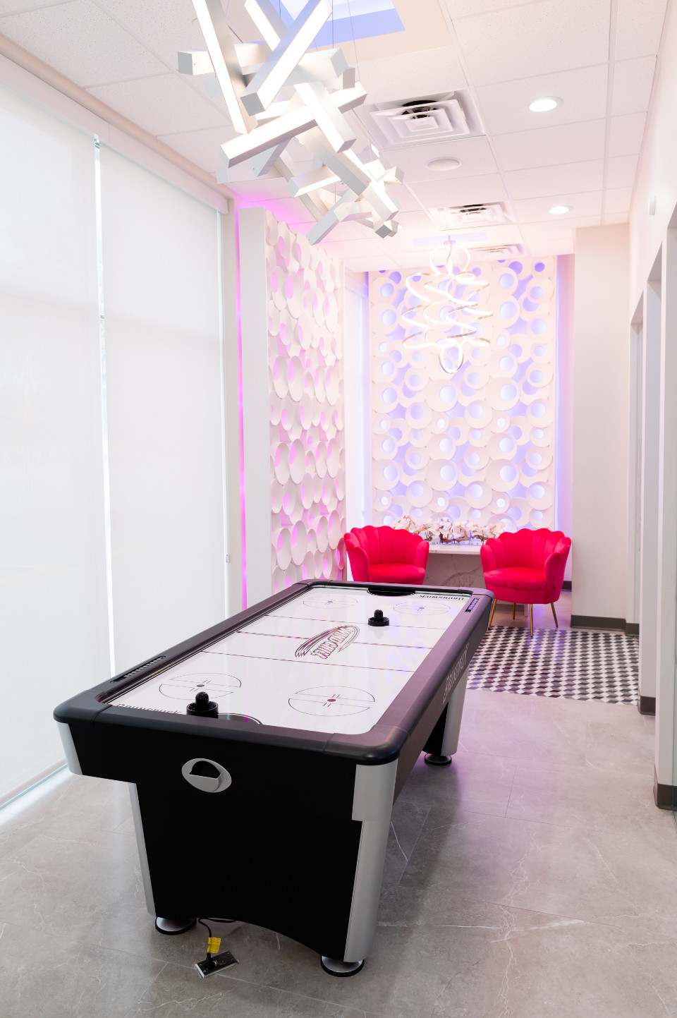A modern game room with an air hockey table in the foreground, two pink chairs, a small table, and decorative white wall panels with purple lighting in the background.