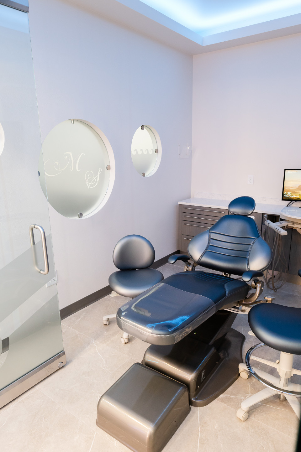 A modern dental office with a blue dental chair, two stools, a computer on a desk, and frosted round windows.