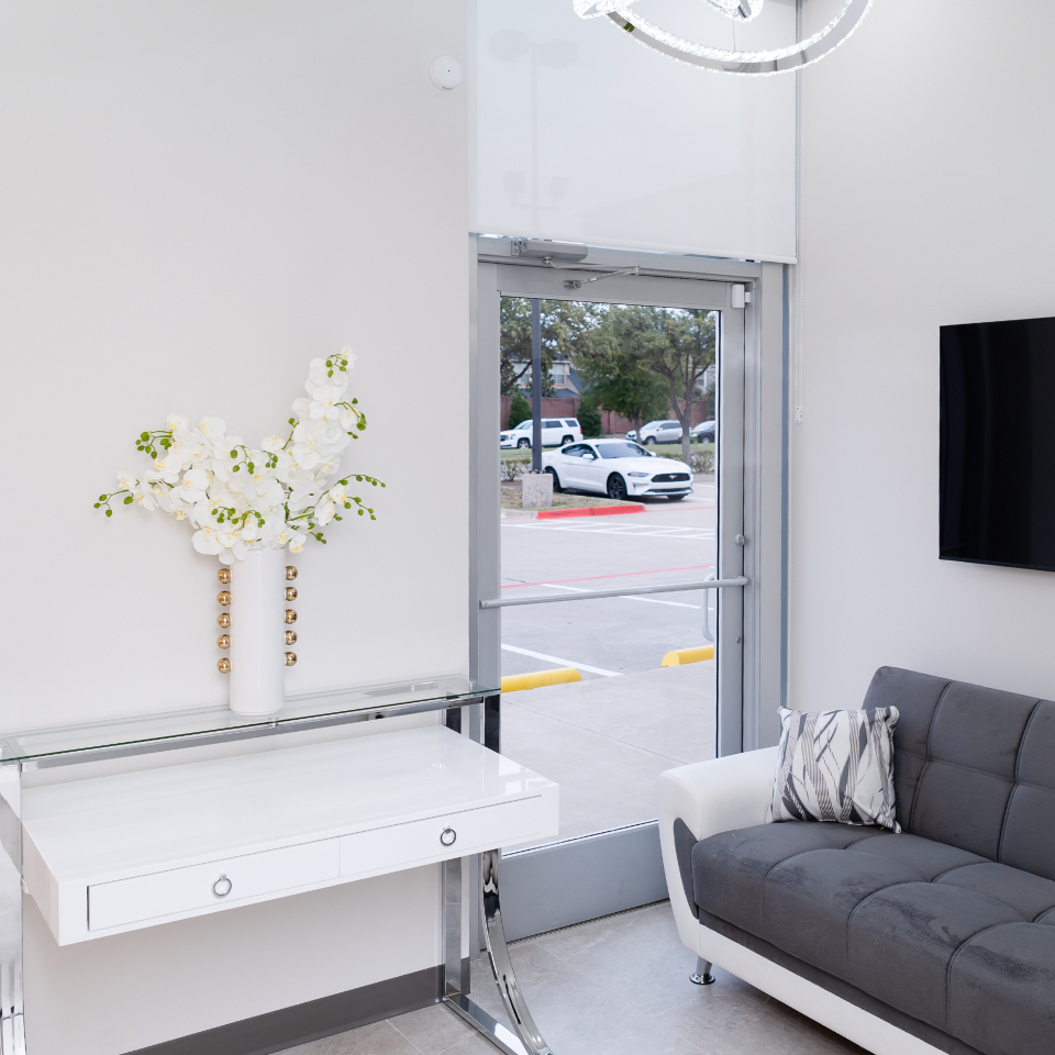 A modern waiting room with a gray couch, a glass table holding a white vase of flowers, and a flat-screen TV; a glass door reveals parked cars outside.