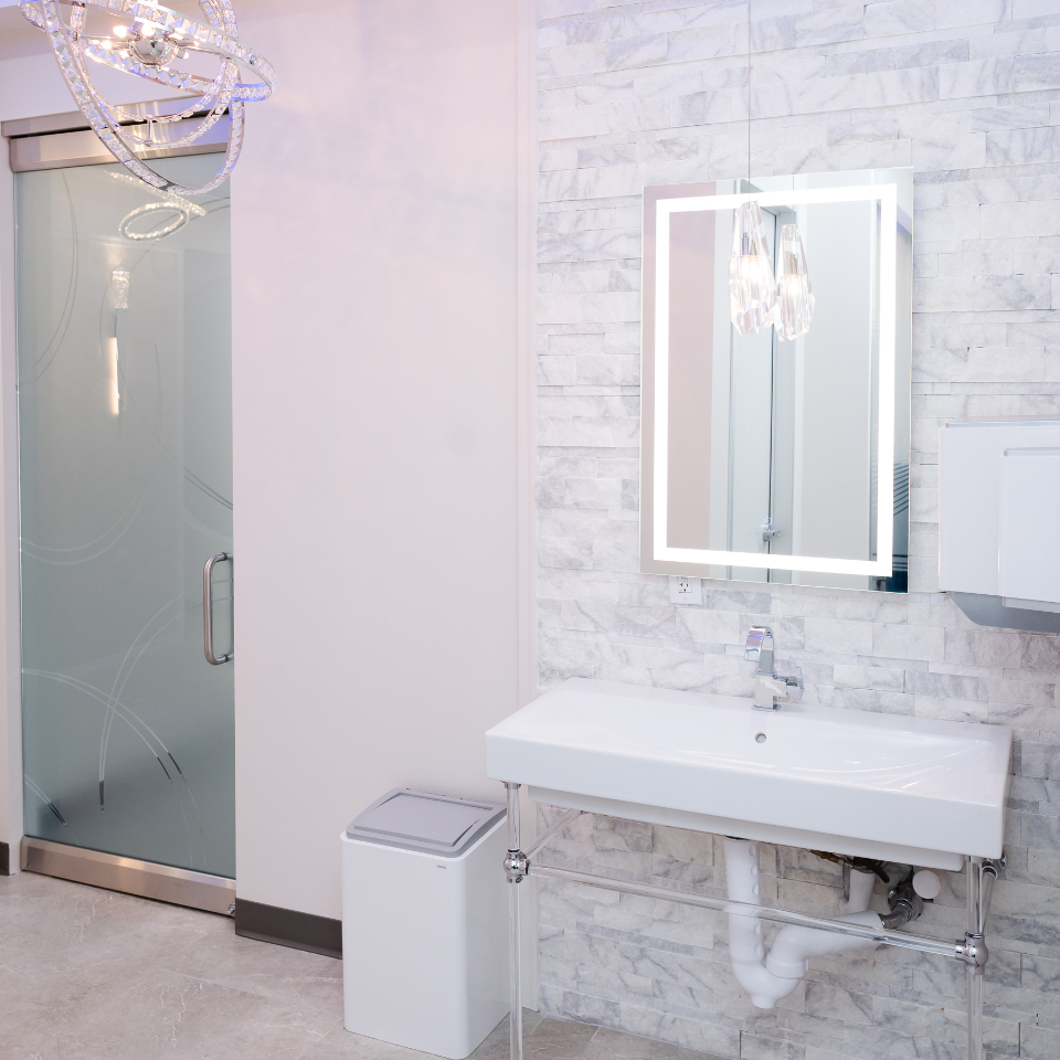 Modern bathroom with a frosted glass door, rectangular sink, illuminated mirror, and white tile walls. A trash bin is placed beside the sink.