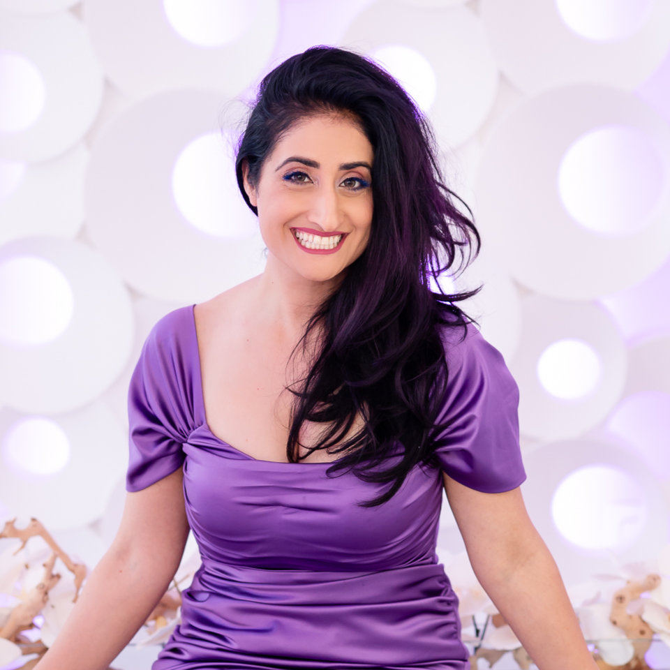 A woman with long dark hair wearing a shiny purple dress sits and smiles in front of a white circular patterned background.