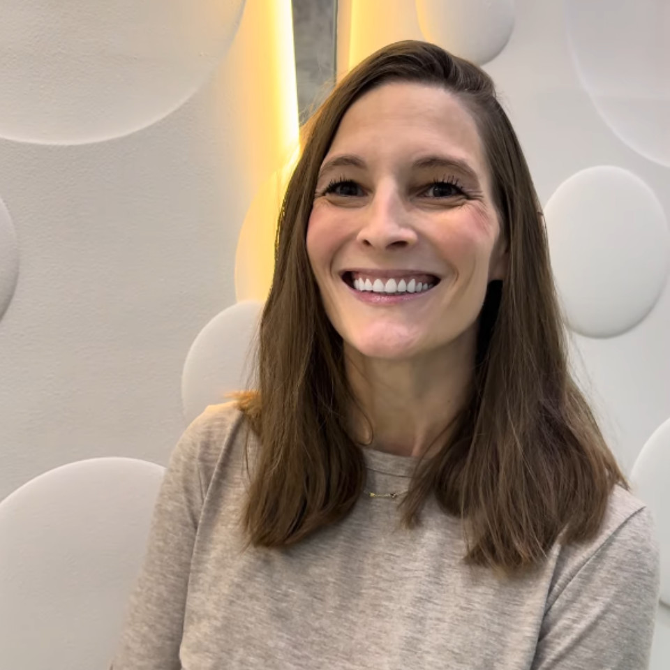 A woman with long brown hair, wearing a beige top, smiles at the camera against a white textured wall with circular patterns and yellow lighting.