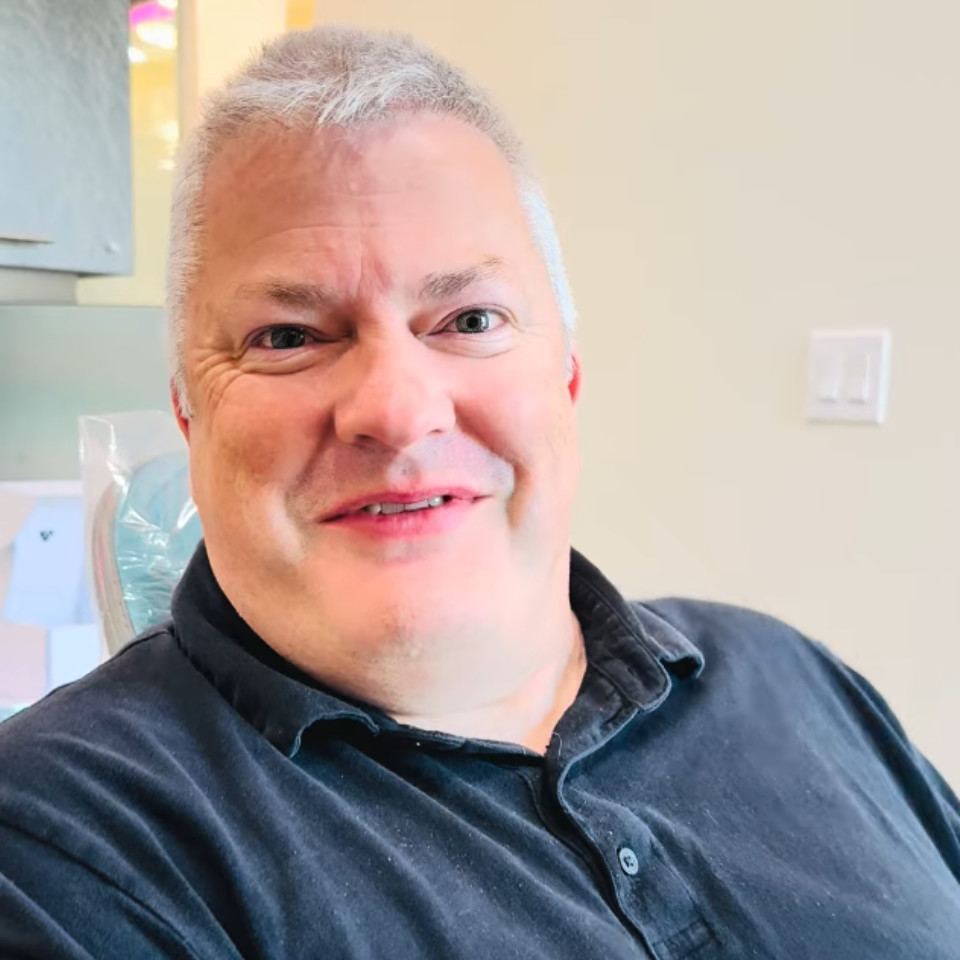 A middle-aged man with short gray hair, wearing a dark polo shirt, sits indoors and smiles at the camera.