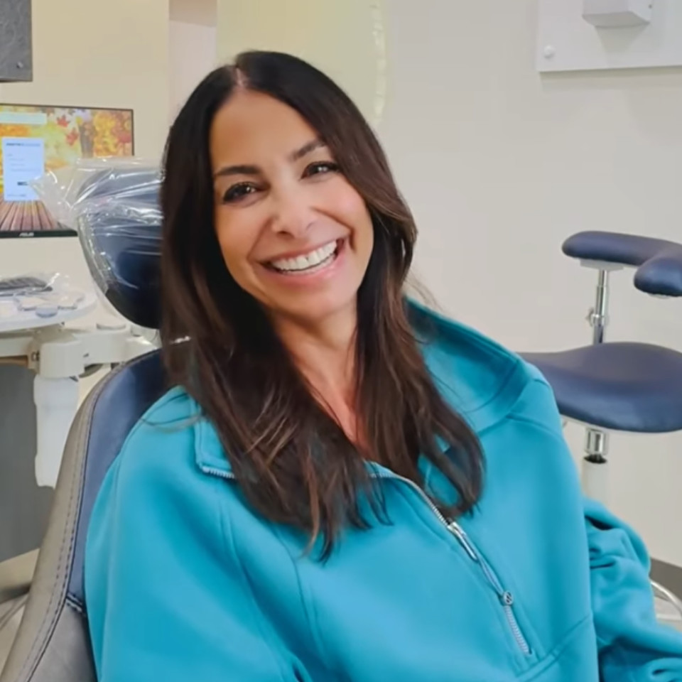 A woman with long brown hair wearing a teal jacket smiles while sitting in a dental chair in a clinic.