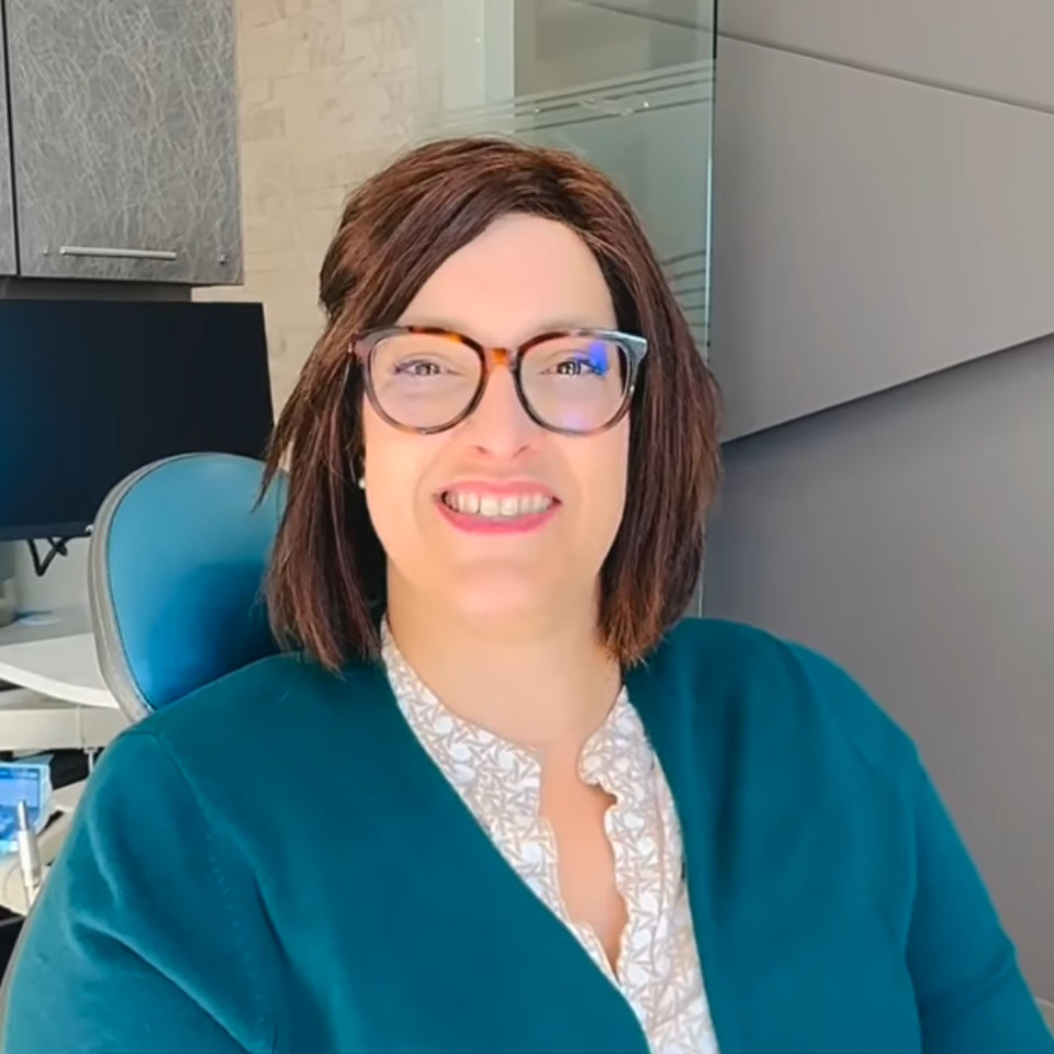 A person with short brown hair and glasses, wearing a teal cardigan and patterned blouse, sits and smiles in a modern office setting.