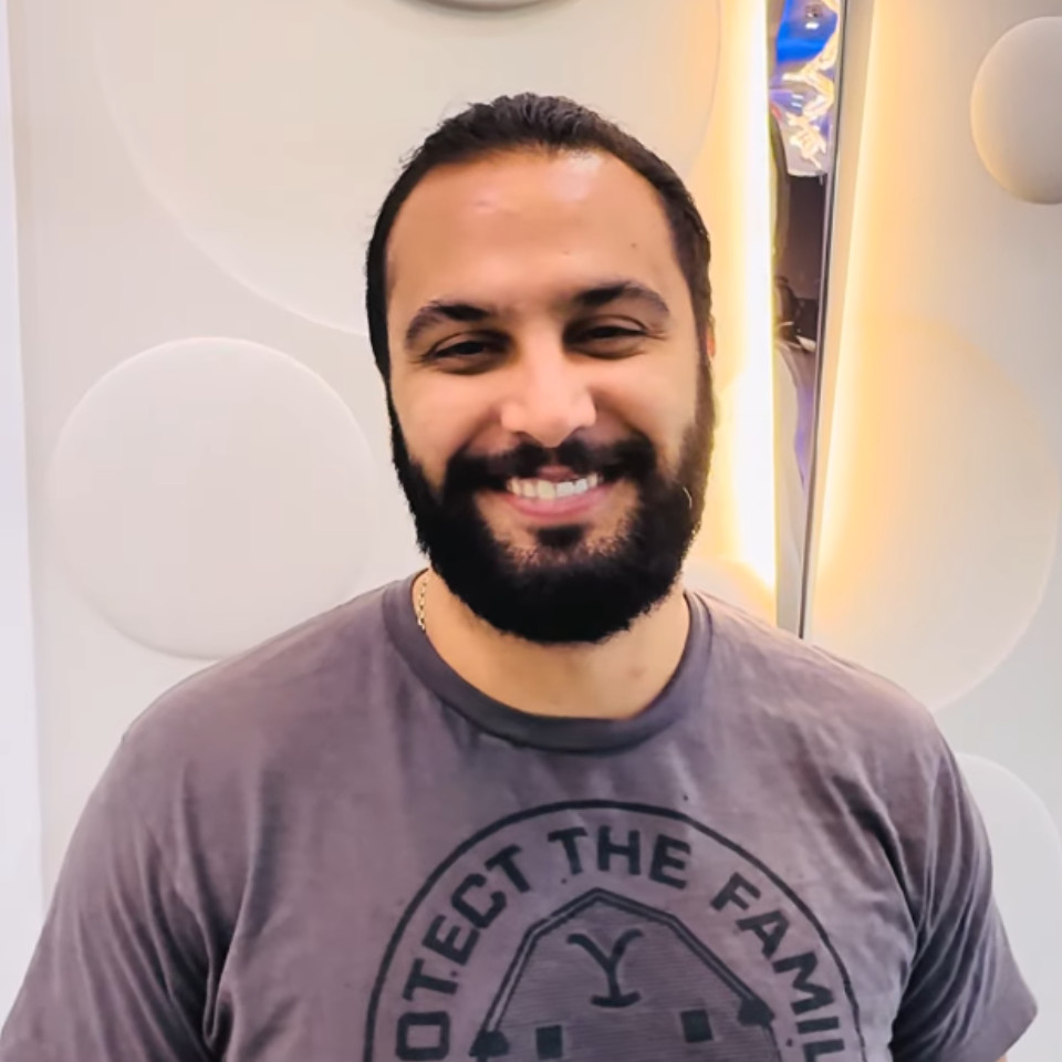 A man with dark hair and a beard smiles at the camera, wearing a grey t-shirt with a circular design that reads "PROTECT THE FAMILY" on the front.