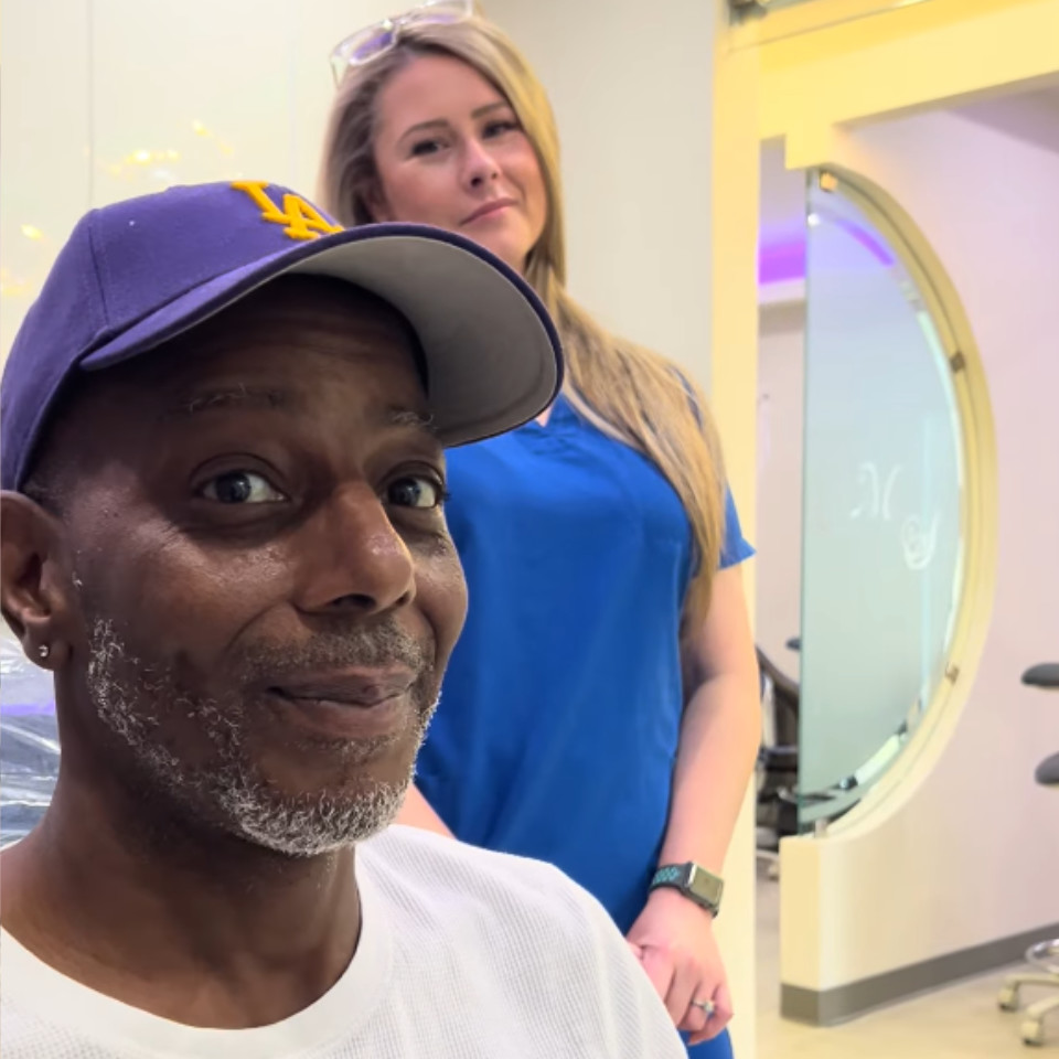 A man in a blue LA cap and white shirt sits in a salon chair, while a woman in blue scrubs stands behind him, both looking at the camera.