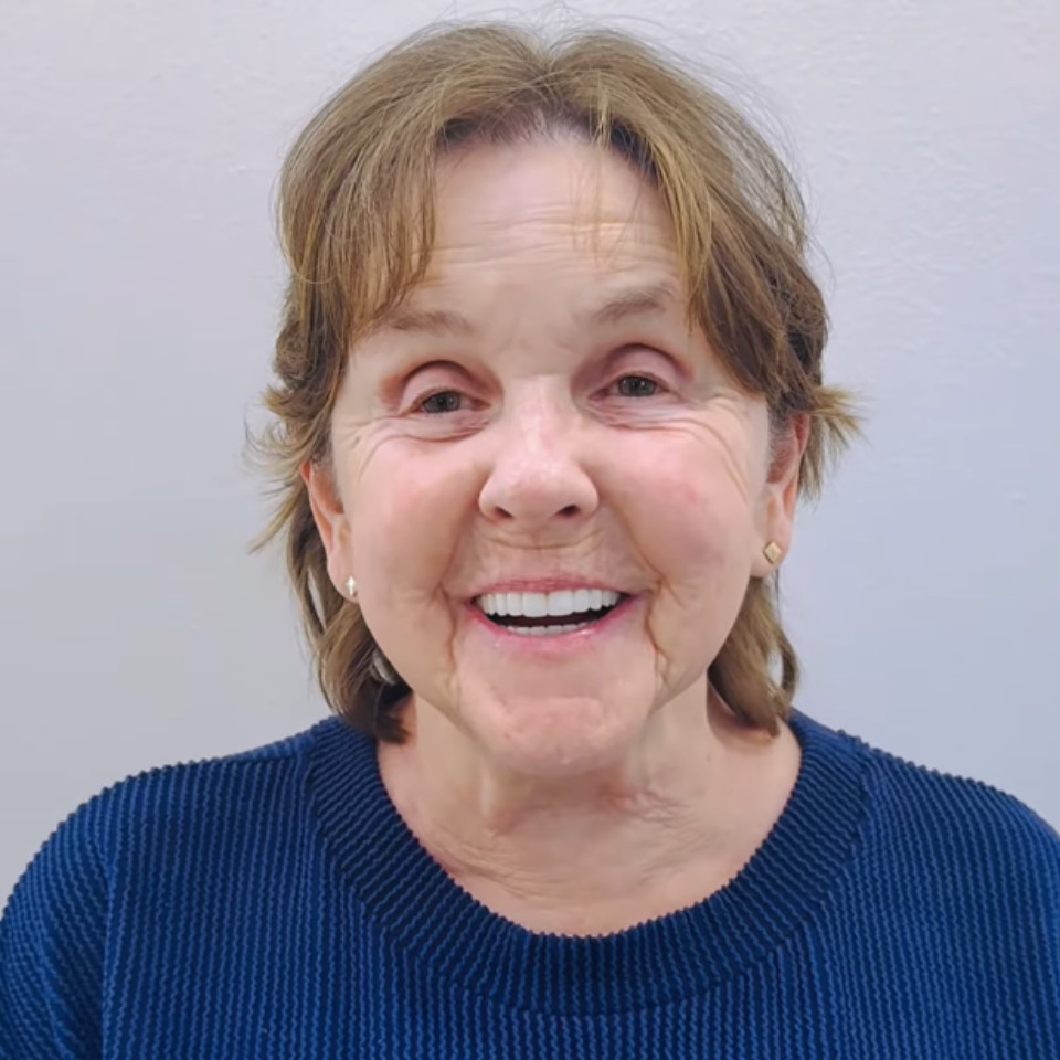 An older woman with light brown hair and a dark blue sweater smiles in front of a plain light-colored wall.