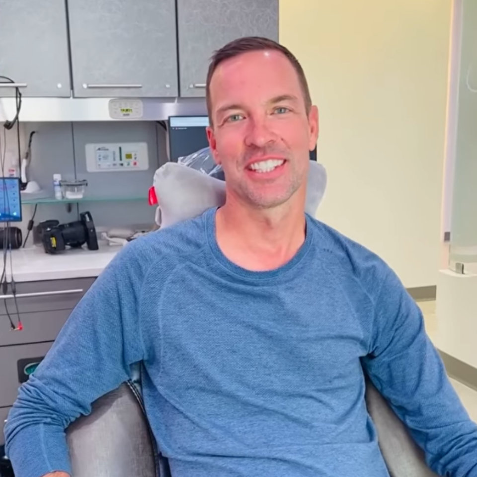 A man in a blue long-sleeve shirt sits and smiles in a modern dental or medical office.