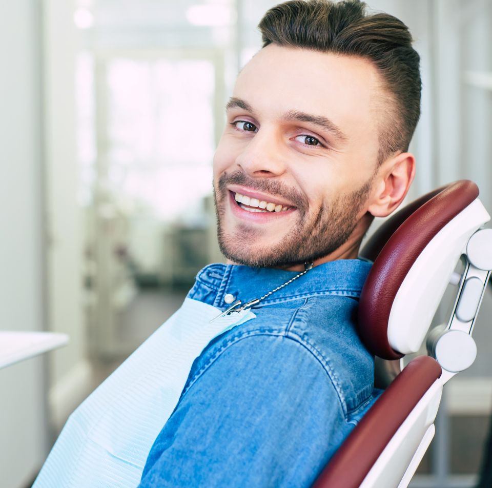 A man in a denim shirt sits and smiles in a dental chair, wearing a dental bib, inside a brightly lit dental office.