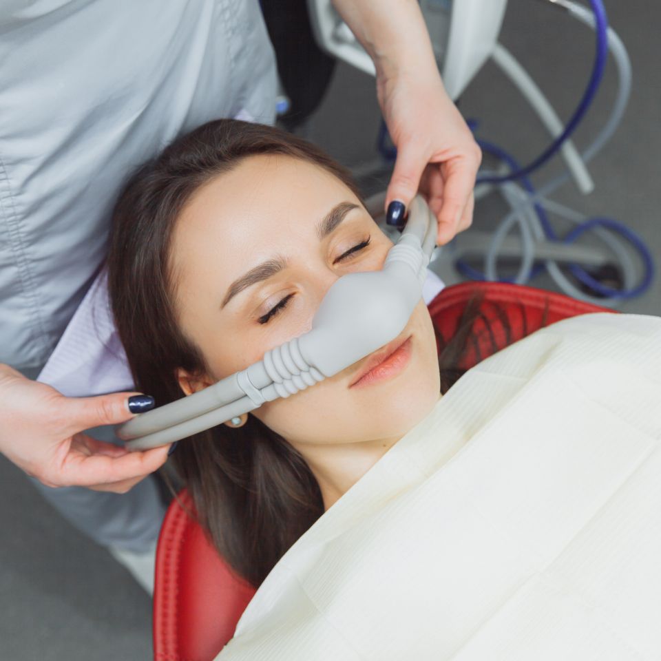 A woman reclines in a dental chair with her eyes closed while a dental professional fits a sedation mask on her face.