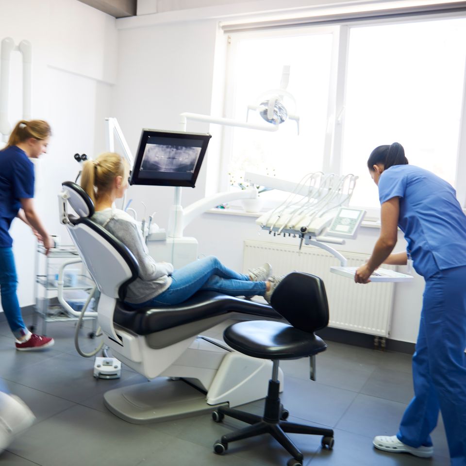 A person sits in a dental chair while two dental professionals in scrubs prepare equipment in a brightly lit dental clinic.