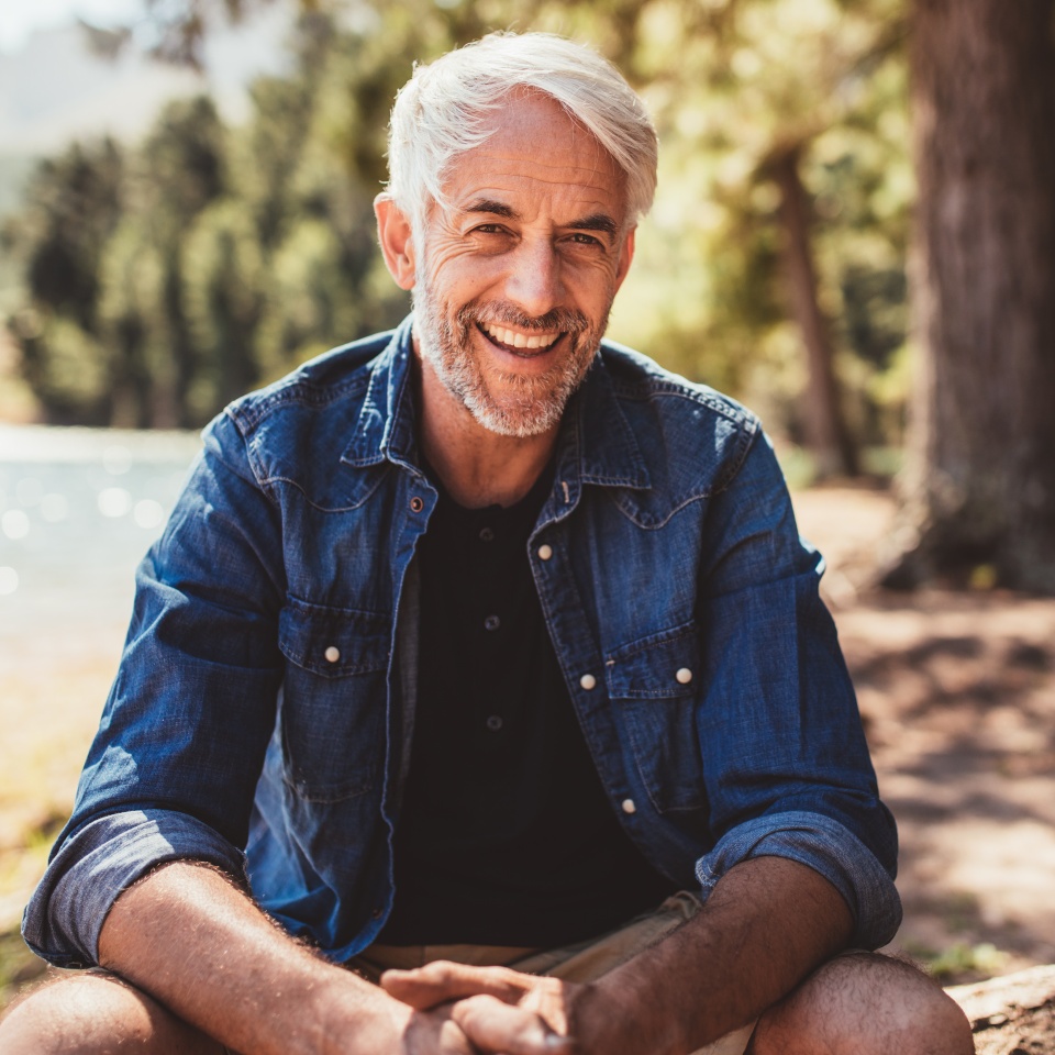 Portrait of happy mature man sitting near a lake looking at camera and smiling. Senior caucasian man sitting on a log by the lake on a summer day.